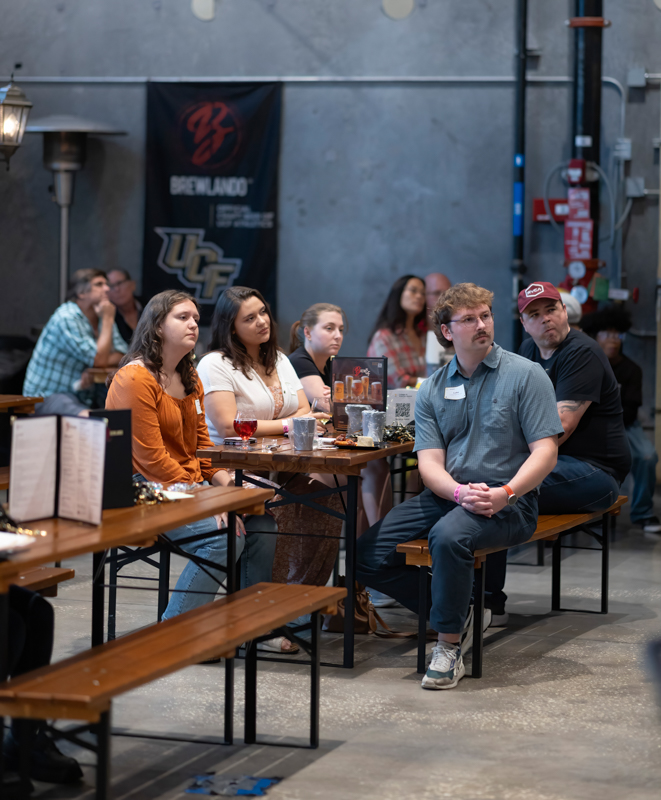 Group of adults seated at long wooden tables in a gray industrial room, attentively watching something off-camera; banners with logos on the wall behind them