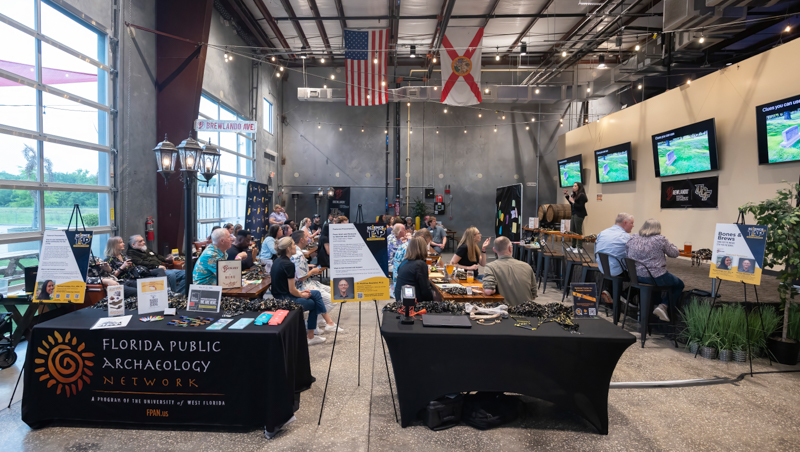 Audience seated at tables in an industrial-style venue listening to a presenter; flags hang overhead and screens show slides.]','Audience watches a speaker at a Florida Public Archaeology Network event, with exhibit tables and display boards in the foreground.