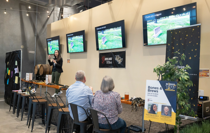 Female speaker presenting to a small audience at a bar-style event; screens behind her show slides.