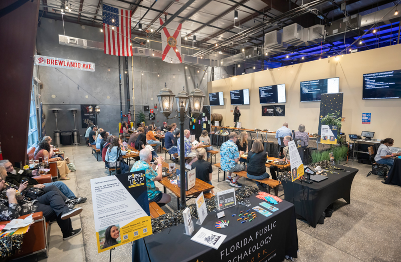 Busy indoor expo with attendees seated at tables, displays, and a presenter on a stage; flags hang from the ceiling.