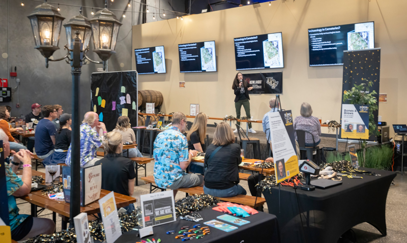 Audience seated at long wooden tables listening to a speaker on a stage in a large event space, with multiple presentation screens behind her and market booths along the sides.