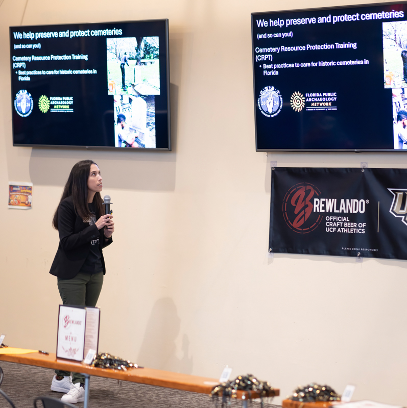 Female presenter with microphone speaks in front of two display screens at a conference or expo event room.