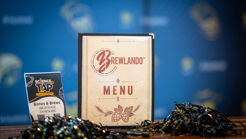 Framed BrewlandO Menu sign with barley hops illustration on a bar, flanked by a blue backdrop; left shows a 'Science on Tap' flyer about Bones & Brews.