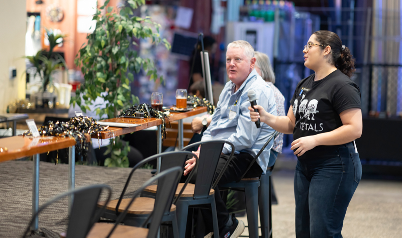 Woman with a microphone interviewing a casually dressed man at a bar-height table during an event, with drinks and festive garland nearby.