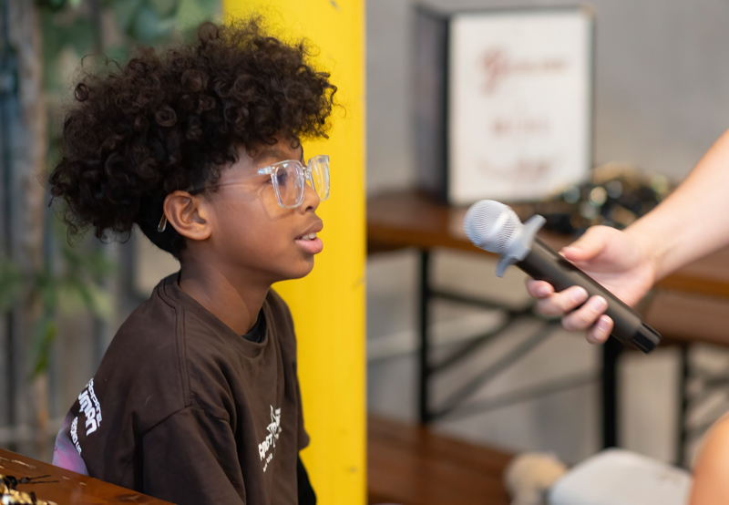 Young person with curly hair and glasses speaks into a handheld microphone during an interview setup in a casual indoor space fell back by a yellow pillar.