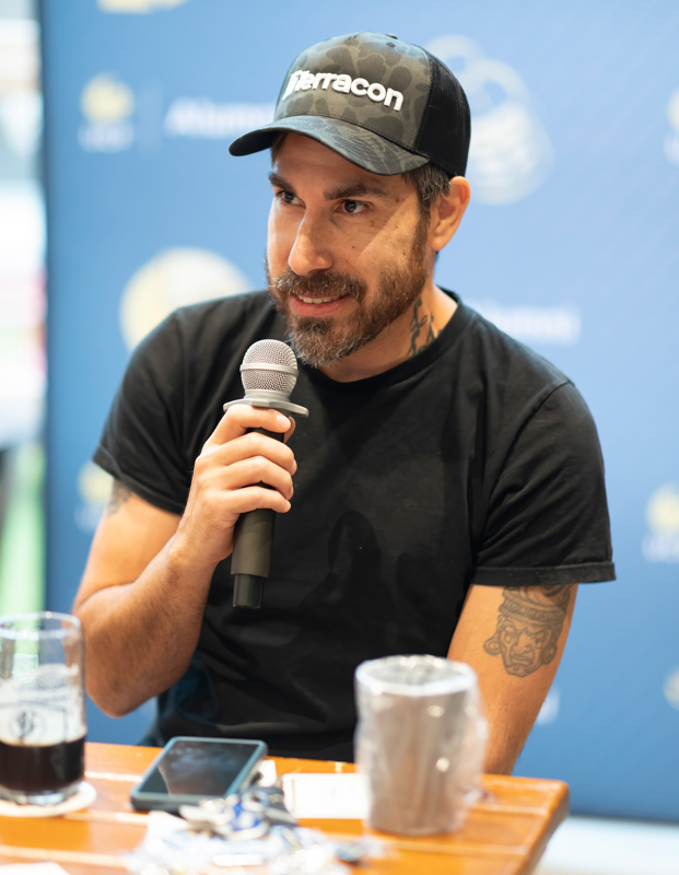 Bearded man in a black shirt and camo Terracon cap speaks into a microphone at a panel, blue backdrop behind him.