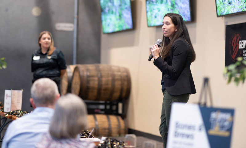 Woman in a black blazer speaks into a handheld microphone on a stage at a wine event, with large wine barrels and screens behind her.