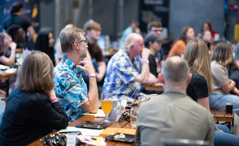 People sit at long communal tables, listening to something off-camera in a casual social gathering, with drinks and snacks on the table and laptops nearby.