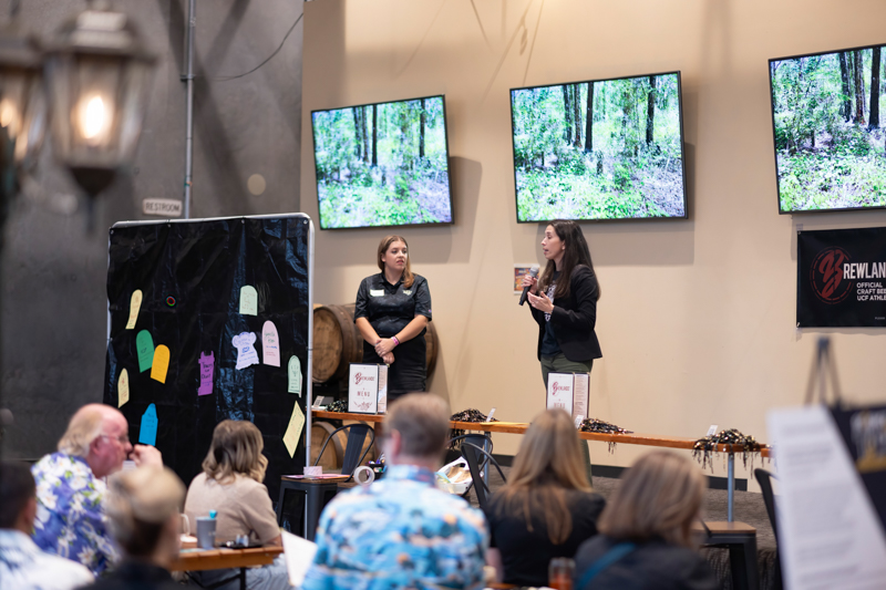 Two women presenting on a stage at a conference with forest images on three screens behind them and an audience watching nearby, indoors.