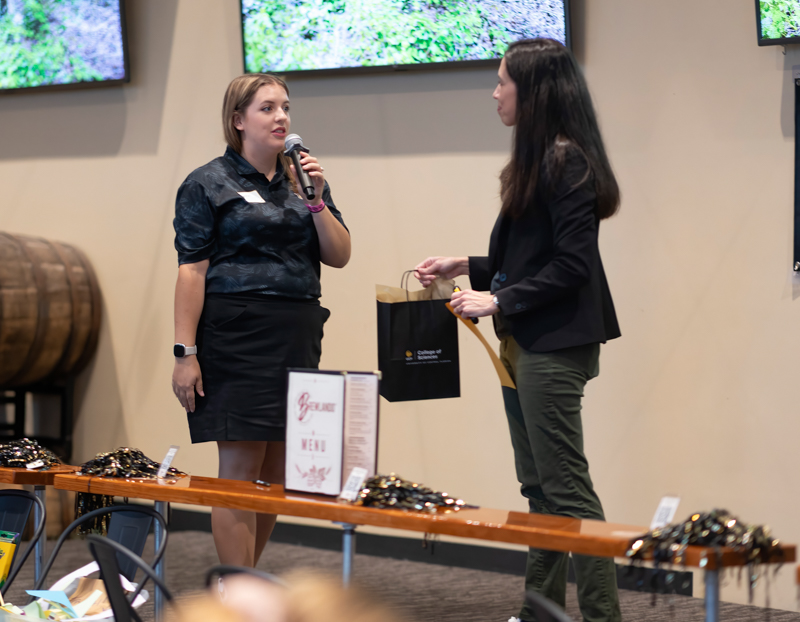 Two women speak at a table-filled event: one holds a microphone while the other hands her a black gift bag.