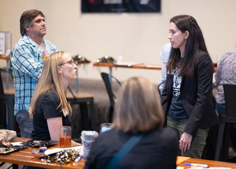 Standing woman in a dark blazer speaks to a seated woman with orange glasses at a workshop-style table, while others listen in the background.