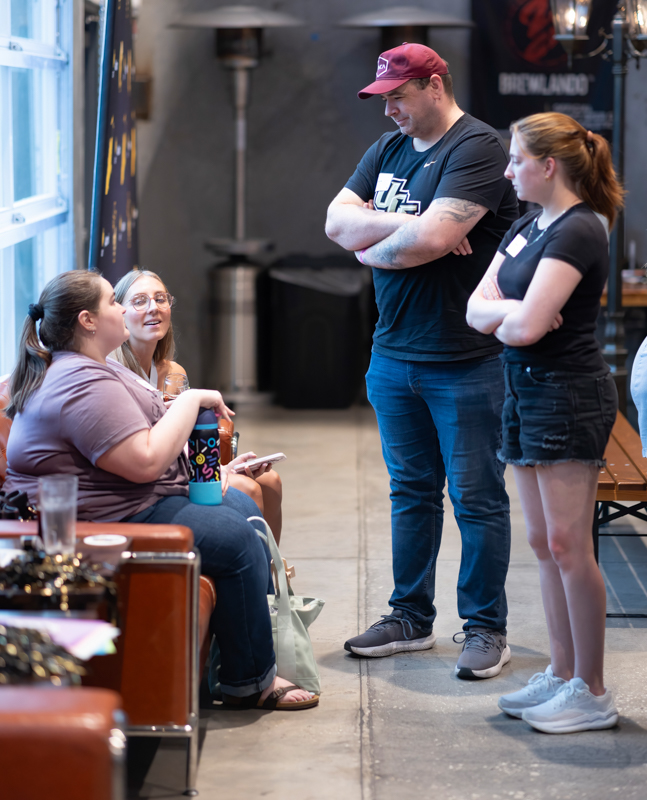 Three adults stand and talk to a seated woman holding a blue tumbler in a casual indoor venue, possibly a social gathering or games area.