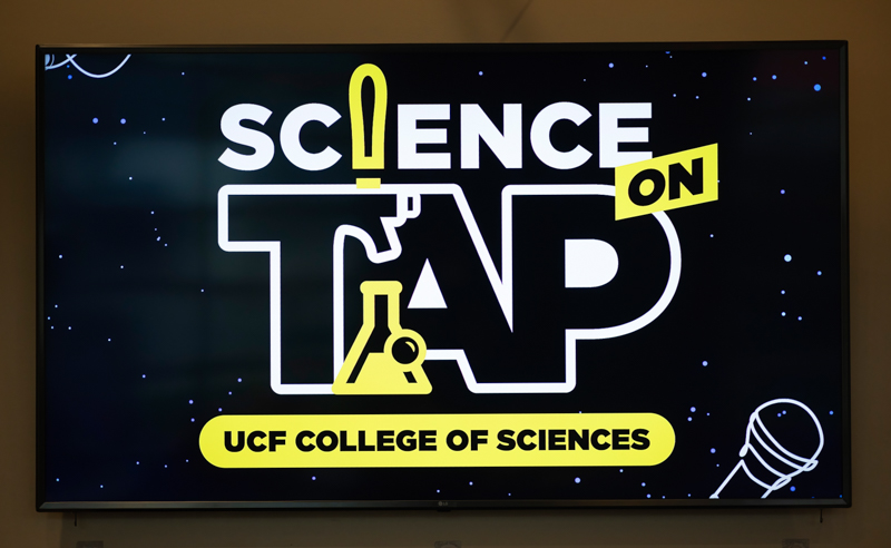 Screen displaying a poster for 'Science On Tap' with the large white 'TAP' letters, yellow accents and a beaker motif, plus a yellow 'UCF College of Sciences' banner and a mic graphic in the bottom-right corner.