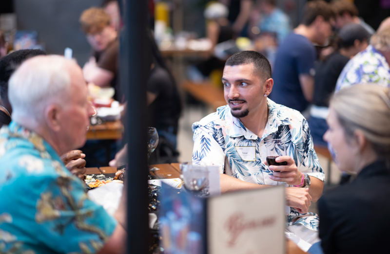 Man in a blue tropical-pattern shirt sits at a communal table, holding a glass of red wine and chatting with others at a social dinner.