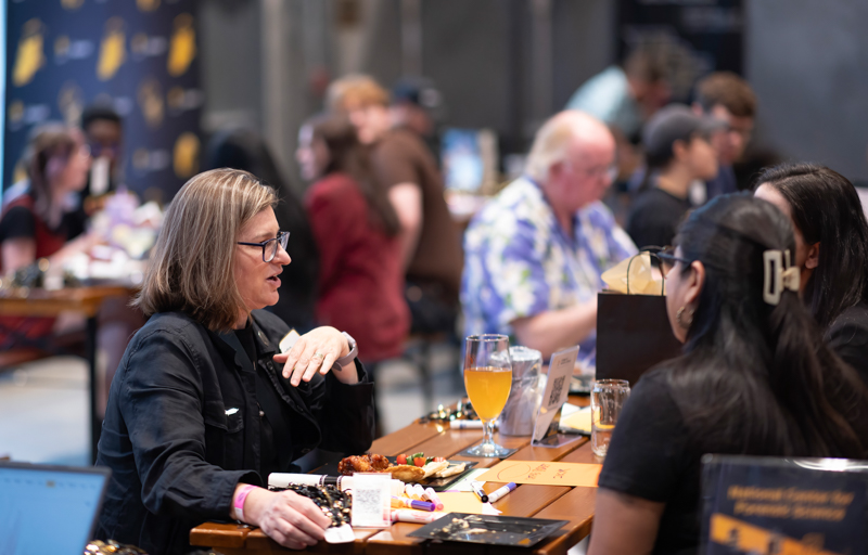 A woman with glasses sits at a wooden table, speaking to another attendee as plates and a glass of orange juice sit nearby at a social event table. The background shows other guests mingling softly blurred.