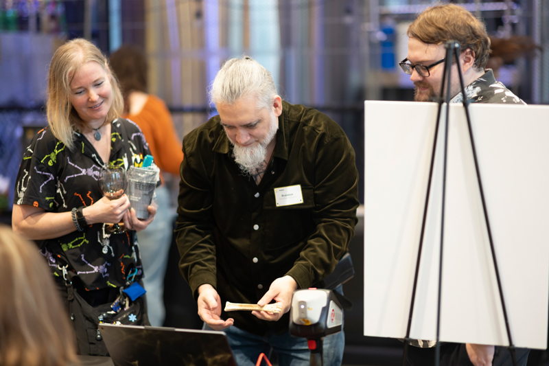 Three people at a conference booth observed a man with a gray beard showing a small wooden piece while a woman holds a glass and a presenter stands by a whiteboard in the background.