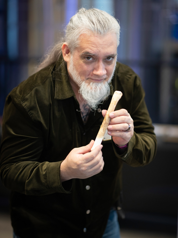 Older man with gray hair and beard leans forward, holding a long bone-like object toward the camera.