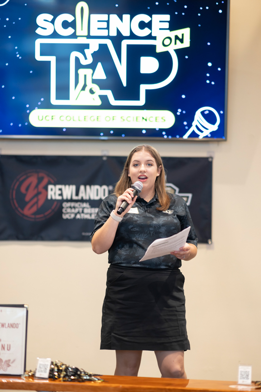 Female presenter on stage at a Science on Tap event, holding a microphone and papers, with a large 'Science on Tap' graphic behind.