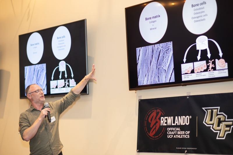 Male presenter with glasses speaks into a microphone, gesturing toward slides about bone biology projected on two screens.