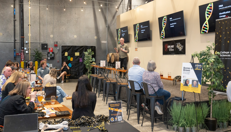 Presenter speaks to an audience in a brewery-style venue; DNA slides on screens behind, attendees at long tables with drinks and laptops.