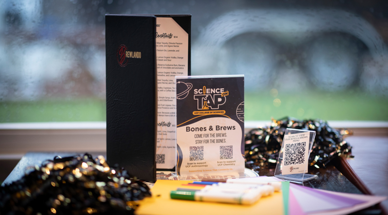 Promotional display for a Science Tap Bones & Brews event with a black vendor box, flyer, and QR code sign on a windowsill table.