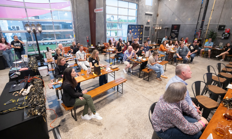 Audience seated at wooden benches in a large industrial-style venue, listening to a talk or presentation near a small stage with holiday lights.