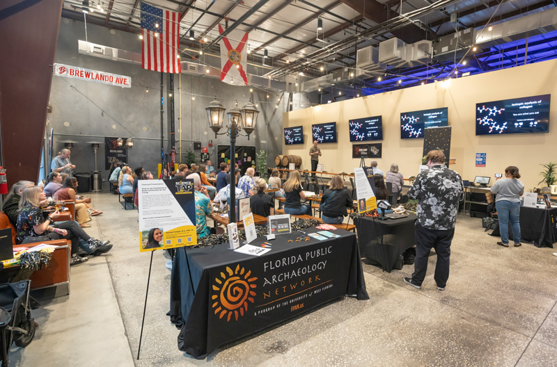 Visitors attend an indoor expo around a Florida Public Archaeology Network booth with informational posters on tables and staff at a long black table.