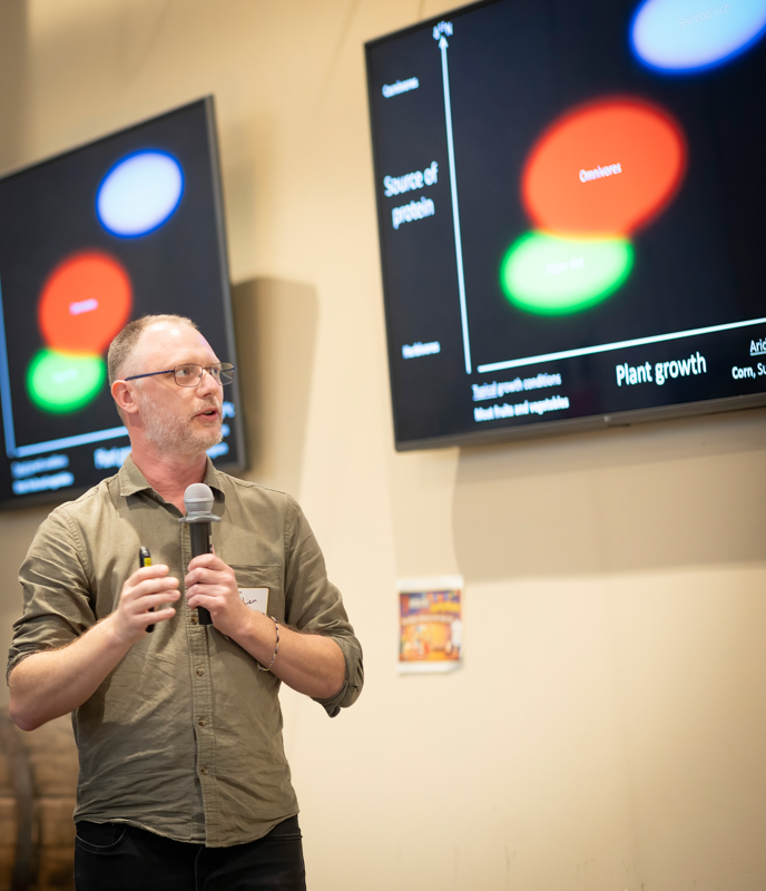 Man wearing a khaki shirt speaks into a handheld microphone during a presentation, with large screens showing a bubble diagram chart in the background.