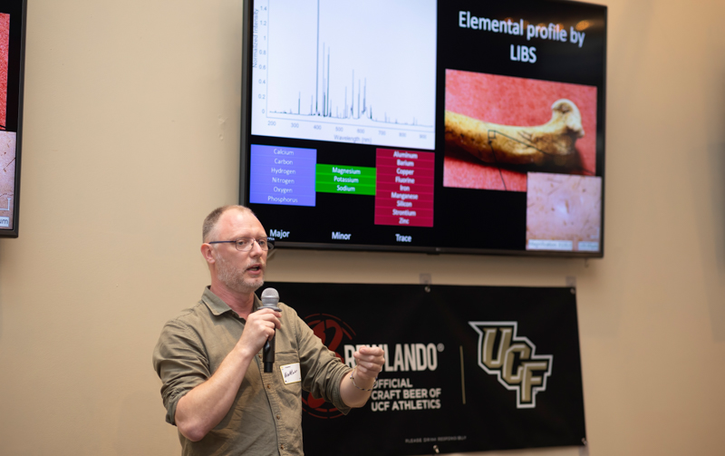 Male presenter speaks into a microphone in front of a large screen displaying charts and the 'Elemental profile by LIBS' slide.
