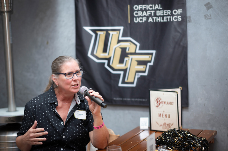 Woman speaking into a handheld microphone at a table, with a UC F banner reading 'Official Craft Beer of UCF Athletics' in the background.