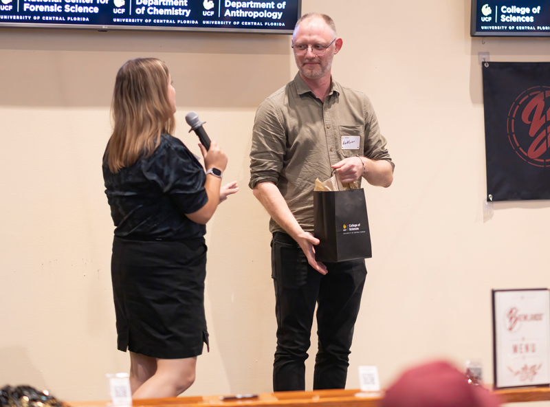 Woman with microphone presents an award or gift bag to a man on a stage during a campus event.