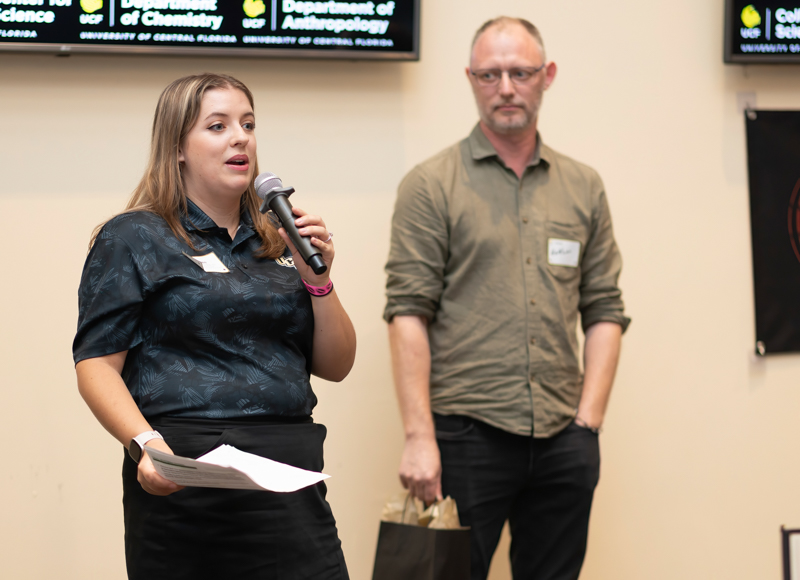 Woman speaks into a microphone, holding papers, at an event; a man in a green shirt stands in the background with a bag nearby.