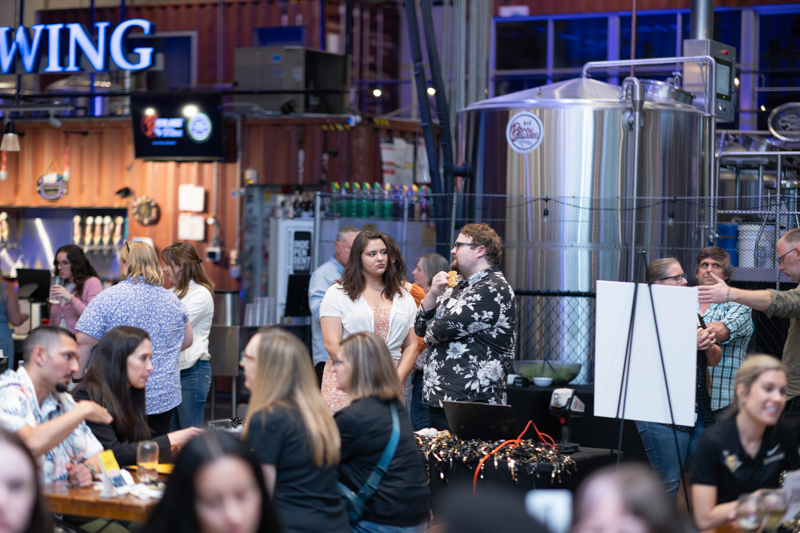 People socialize at a brewery event, with stainless beer tanks and a bar in the background.