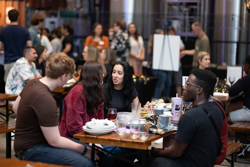 Group of friends chatting at a long communal table during a casual social event with plates and drinks nearby.