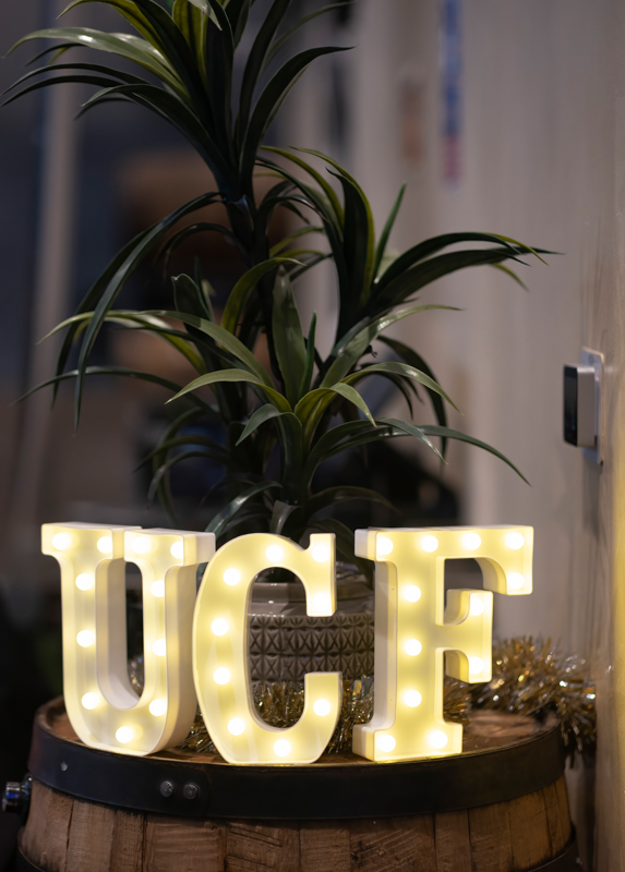 Illuminated UCF letters on a wooden barrel planter with a tall green plant in the background.