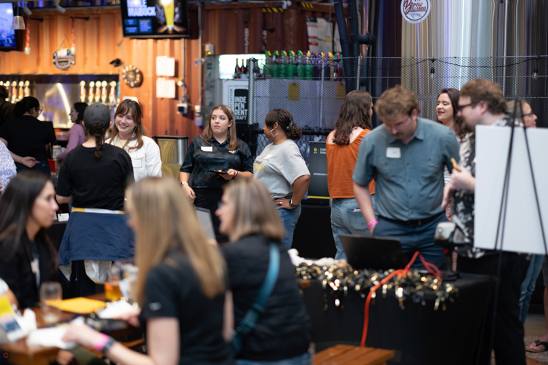 People mingle at a social event with booths and jewelry on display, in a crowded indoor venue with drinks in the background.