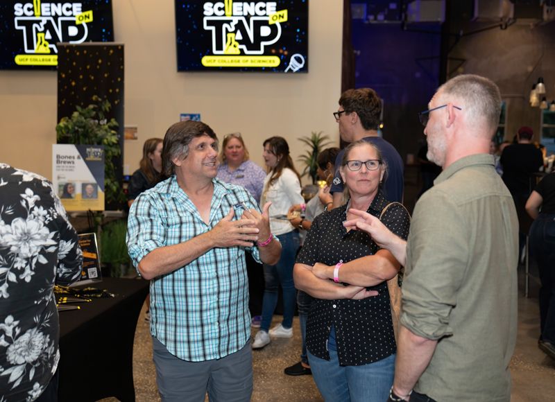 Group of adults conversing at a Science on Tap event, with screens showing the event logo in the background.
