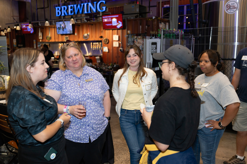 Group of five people standing in a brewery taproom, chatting and holding drinks.