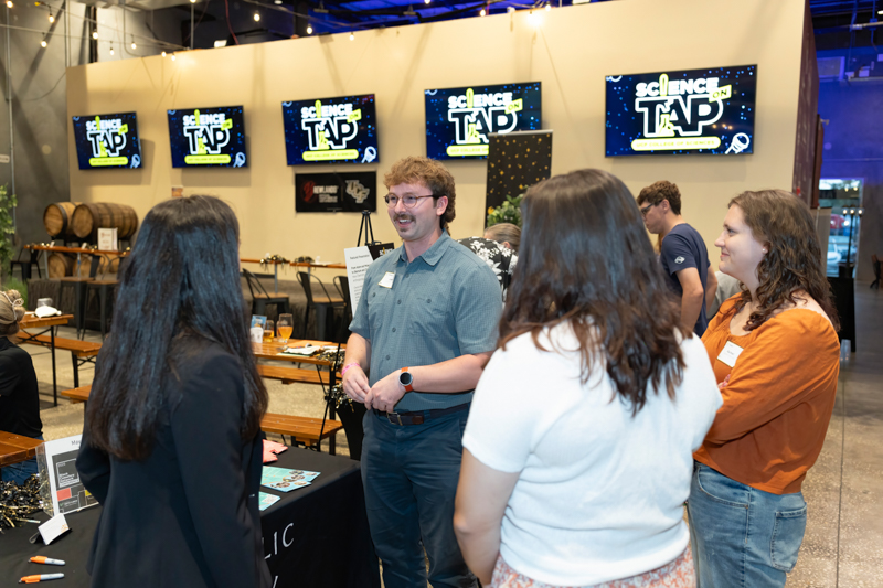 Group of four people chatting at a science event booth, with 'Science on Tap' screens in the background.