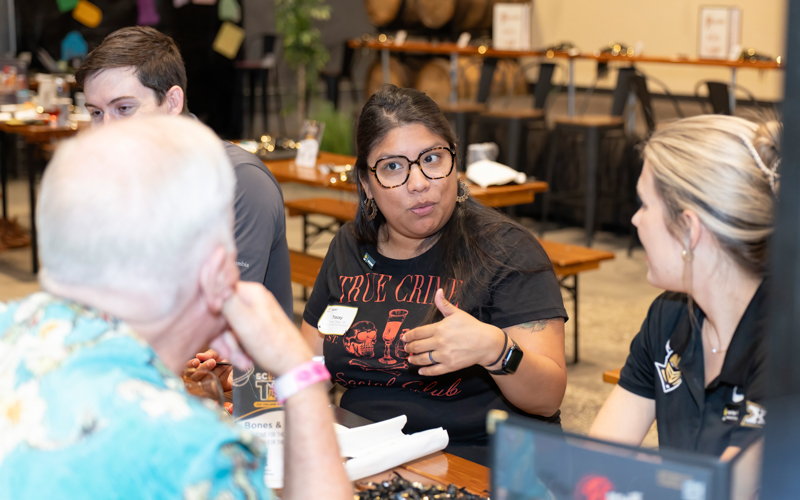 Woman with glasses explains something to a group at a table during an event, others listening attentively.
