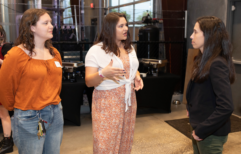 Three women engage in conversation at a casual indoor event, one gesturing as she speaks to the others.