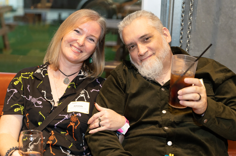 Smiling couple seated together at a casual gathering; woman wears a skeleton-pattern shirt and a name tag reading Amanda, holding a drink while the man beside her smiles with a beard and dark shirt.