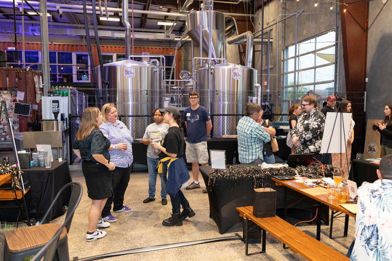 Group of people chatting near shiny stainless steel brewing tanks during a brewery open house.