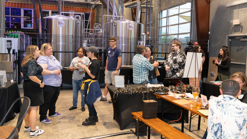 Group of people chatting and tasting drinks at a brewery event, with large stainless fermentation tanks visible in the background.