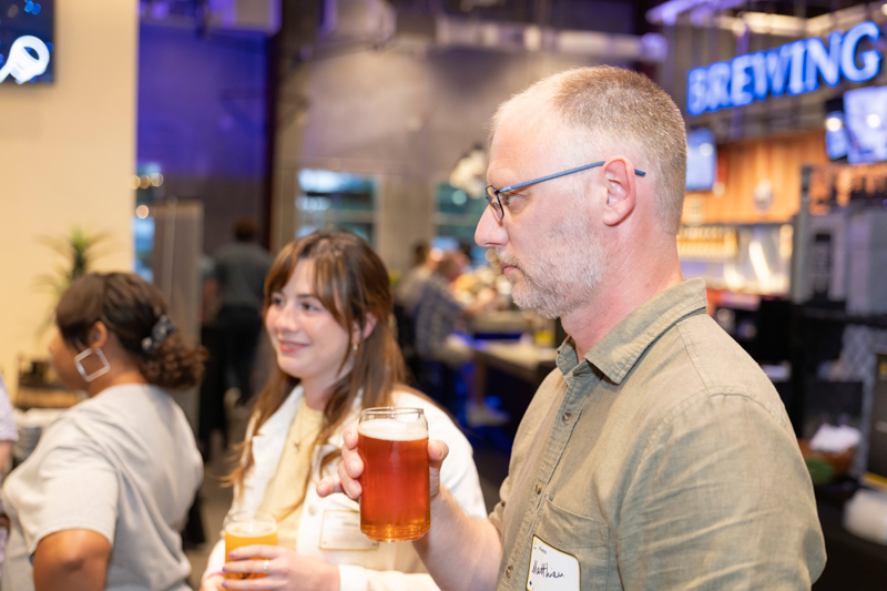 Man with glasses holding a pint of amber beer at a brewery bar, woman beside him smiling with another glass, background patrons.