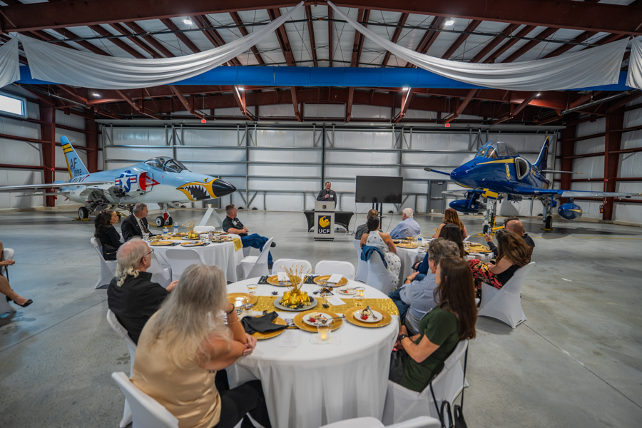 Guests dine at round tables in a hangar, listening to a speaker at a podium with two jets on display behind them, festive decorations overhead.