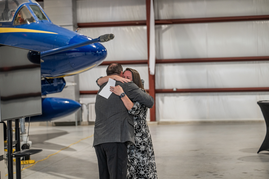 Two people hug in a hangar beside a blue aircraft, one holding a white folder.