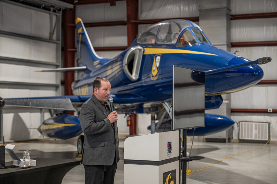 Man in a gray blazer speaks into a microphone at a podium in an aviation hangar, with a blue and yellow jet clearly visible behind him.