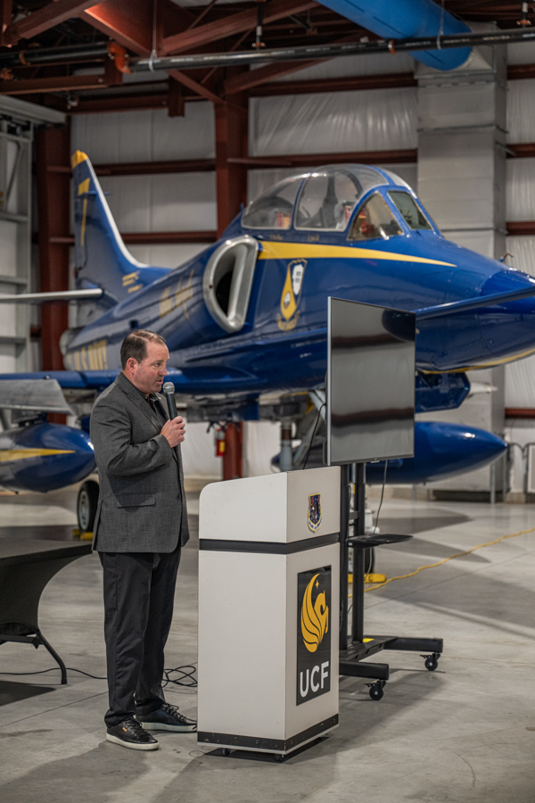 Man in a gray blazer speaks into a microphone at a podium with the UCF logo, set in a hangar beside a blue aircraft.