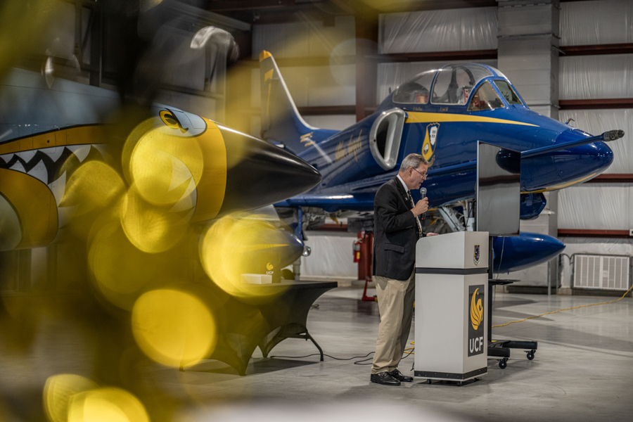 Man in a blazer speaks at a podium inside a hangar, with a blue and gold aircraft on display behind him amid yellow bokeh.